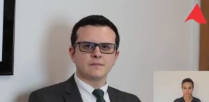 Imagem mostra um trecho do vídeo apresentado pelo juiz de Direito Bruno Cortez Torres Castelo Branco. Ele veste terno escuro, camisa branca, e está numa sala com parede branca e um detalhe de um quadro ao fundo
