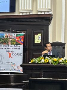 A foto mostra o desembargador em uma mesa de madeira ornada com flores. Ele usa terno escuro, gravata roxa, e fala ao microfone. Ao lado há um banner com o nome do Cojud