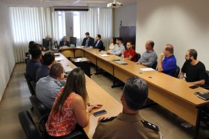 A foto mostra o público presente na audiência pública, entre homens e mulheres. Eles estão sentados em uma mesa de reunião de madeira. As paredes da sala são brancas e ao fundo há uma janela ampla com cortinas claras