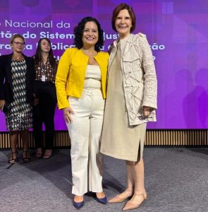 A imagem mostra quatro mulheres em um palco. Em primeiro plano está a juíza de Direito substituta do Juízo Militar de Minas Gerais Renata Rodrigues de Pádua de calça e blusa bege, blazer amarelo e sapato azul. Ela tem cabelos enrolado na altura dos ombros e sorri para a foto. Ao seu lado está a presidente do STM, ministra Maria Elizabeth Rocha, de vestido e casaco bege, e sapato caramelo. Ela também sorri. Ao fundo estão outras duas mulheres e um telão sobre um painel de madeira