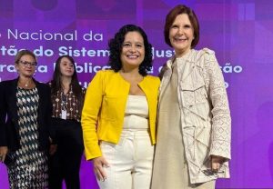 A imagem mostra quatro mulheres em um palco. Em primeiro plano está a juíza de Direito substituta do Juízo Militar de Minas Gerais Renata Rodrigues de Pádua de calça e blusa bege e blazer amarelo. Ela tem cabelos enrolado na altura dos ombros e sorri para a foto. Ao seu lado está a presidente do STM, ministra Maria Elizabeth Rocha, de vestido e casaco bege. Ela também sorri. Ao fundo estão outras duas mulheres e um telão