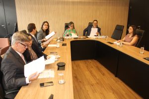 A imagem mostra três homens e quatro mulheres sentados em uma mesa de reunião de madeira com detalhes em preto, mesmas cores da parede da sala. Eles mexem em papéis e conversam entre si