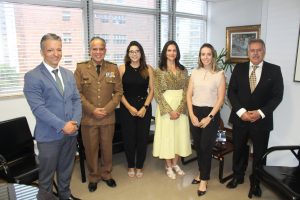 A foto mostra seis pessoas. O secretário especial da Presidência vestindo terno cinza; o vice-presidente, desembargador James Ferreira Santos, usa farda marrom da Polícia Militar de Minas Gerais; a nova oficial judiciária veste blusa e calça preta, e usa óculos; a diretora da DRH veste saia amarela e blusa estampada; a gerente de Secretaria da 1ª AJME usa calça preta e blusa clara; e o assessor jurídico veste terno preto com gravata xadrez. Eles estão de pé em uma sala com uma ampla janela ao fundo e um quadro de flores na parede ao lado