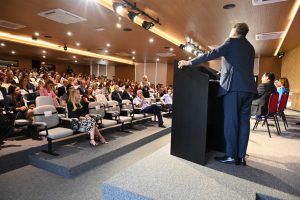 A imagem mostra um auditório com homens e mulheres sentados na plateia diante de um palco, onde estão duas mulheres e um homem sentados, e mais um homem em um púlpito.