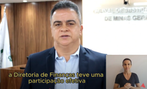 A imagem mostra trecho do vídeo apresentado pelo gerente da Diretoria Executiva de Finanças, Ângelo de Magalhães, que veste terno azul escuro e camisa azul claro. Ele está em pé na sala do Tribunal Pleno do TJMMG. Ao fundo, à esquerda há três bandeiras e um letreiro com o nome do TJMMG. No canto inferior direito há uma intérprete de libras usando camisa preta.