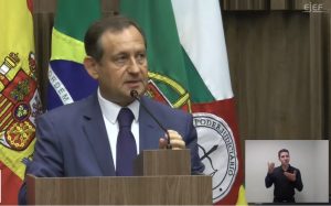 A foto mostra o presidente do TJMG, desembargador Luiz Carlos Corrêa Junior. falando ao microfone atrás de um púlpito de madeira. Ele veste terno azul com gravata da mesma cor. Ao fundo há quatro bandeiras e no canto inferior direito há um intérprete de libras usando camiseta preta.