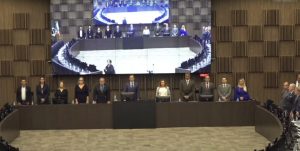 A imagem mostra a composição do dispositivo de honra na abertura do 1º Simpósio Internacional de Justiça Terapêutica, com 11 pessoas, entre homens e mulheres. O vice-presidente do TJMMG, desembargador James Ferreira Santos é o antepenúltimo da esquerda para a direita e está usando terno marrom com gravata vermelha. Ao fundo há uma parede marrom e um telão com a transmissão ao vivo do evento