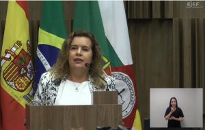 A imagem mostra a reitora da UFMG, Sandra Regina Goulart Almeida, falando ao microfone atrás de um púlpito de madeira. Ela veste uma blusa branca e um blazer branco com detalhes em preto. Ao fundo há quatro bandeiras e no canto inferior direito há uma intérprete de libras usando camiseta preta