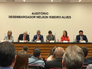 A imagem mostra várias pessoas, entre homens e mulheres, participando do Seminário da Nova Política Nacional de Segurança Institucional do Poder Judiciário. Há seis pessoas em destaque compondo a mesa do evento, todos estão sentados atrás de uma mesa de madeira olhando para a plateia que também está sentada. A parede ao fundo é branca e possui um letreiro com os dizeres Auditório desembargador Nelson Ribeiro Alves