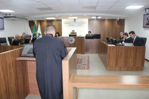 Imagem mostra a composição do Tribunal Pleno. Do lado esquerdo há uma tribuna com dois desembargadores fardados com roupa da PMMG. Ao centro, uma mulher e dois homens, um deles o vice-presidente que presidiu a sessão e estava fardado também com roupa da PMMG. Na tribuna direita três homens, dois deles desembargadores vestindo toga, e um com fardamento do Corpo de Bombeiros. Em primeiro plano, um defensor aparece de costas. Ao fundo há duas bandeiras e uma placa com a logomarca do TJMMG, composta por um triângulo estilizado da bandeira de Minas Gerais e o nome do tribunal por extenso 