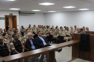 Imagem mostra a plateia presente na sala do Tribunal Pleno do TJMMG, composta por dois civis e 58 militares, entre homens e mulheres fardados. Eles estão sentados em poltronas vermelhas, em uma sala de paredes brancas