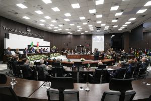 A imagem mostra a Sala de Sessões Ministro Teori Zavascki, do TJSC. Há vários convidados, entre homens e mulheres, a maioria está sentada atrás de mesas longas de madeira. O teto possui várias luzes brancas quadriculadas e as paredes são marrons. No canto direito há uma parede marrom com o letreiro escrito Tribunal de Justiça de Santa Catarina