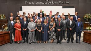 A foto mostra participantes do evento, entre homens e mulheres. Dentre eles está o vice-presidente do TJMMG, desembargador James Ferreira Santos, na primeira fila, vestindo terno preto e gravata vermelha. Ao fundo há uma parede marrom com o letreiro escrito Tribunal de Justiça de Santa Catarina
