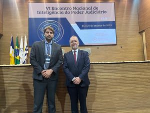 A imagem mostra dois homens de pé, diante de uma bancada de madeira. Eles vestem terno escuro e usam barba. Atrás há quatro bandeiras e um telão com o texto VI Encontro Nacional de Inteligência do Poder Judiciário