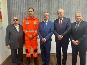 A imagem mostra o novo comandante do 2º BBM vestindo traje laranja dos bombeiros de Minas Gerais. Ele está em uma sala acompanhado por quatro representes do TJMMG. À esquerda está o presidente desembargador Jadir Silva usando terno preto, camisa branca, gravata amarela e óculos escuros. À direita está o desembargador Osmar Duarte Marcelino vestindo terno azul, gravata da mesma cor, camisa branca e usando óculos de grau; o desembargador Rúbio Paulino Coelho usando terno azul, camisa branca e gravata vermelha; e o chefe de gabinete coronel Giovanne Gomes da Silva, de terno e gravata pretos, e camisa branca. Ao fundo há uma parede branca e janela ampla com persiana cinza.