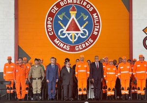 A foto mostra o dispositivo de honra composto por homens e mulheres civis e militares. Na primeira fila há cinco bombeiros vestindo uniforme laranja, um homem vestindo uniforme marrom da PMMG, e os desembargadores do TJMMG de terno. Ao fundo há o brasão do CBMMG