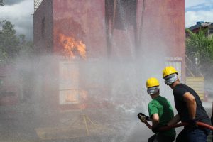 Foto mostra a aula prática de combate à incêndio do 1° Curso de Formação de Brigada de Incêndio do TJMMG, realizado em janeiro de 2023. Em primeiro plano, no canto direito, dois homens aparecem de costas, segurando a mangueira de incêndio. Um está de camiseta verde, outro de camiseta preta, ambos estão com um capacete amarelo. A mangueira joga água em uma edificação de fachada vermelha, onde há uma estrutura pegando fogo
