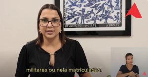 A imagem mostra a juíza de Direito substituta do Juízo Militar Carolina Aleixo Benetti de Oliveira Rodrigues usando camisa preta e óculos. A sala está decorada com uma planta e um quadro com a representação de pássaros azuis. No canto superior direito há um detalhe da logomarca do TJMMG e no canto inferior há uma janela com a intérprete de libras vestindo preto