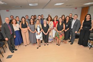A foto mostra um grupo de pessoas, entre homens e mulheres, em uma sala. Eles estão de pé. Na primeira fila, de vestido branco e casaco preto, está Tatiana Reis, gerente da Ascom/TJMMG