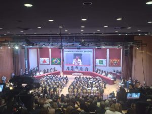 A imagem mostra a sala de um teatro, com pessoas, entre homens e mulheres, sentadas na plateia e no palco. Ao fundo há uma projeção com imagens da transmissão do próprio evento