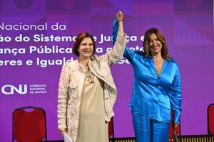 A imagem mostra duas mulheres em um palco, sorrido de mãos dadas e erguidas. À esquerda está a presidente do STM, de vestido e casaco bege. À direita está a conselheira do CNJ de conjunto de camisa e calça azul