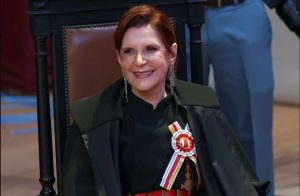 A imagem mostra a ministra Maria Elizabeth Rocha sentada em uma cadeira. Ela está de cabeos presos, usa roupa e toga pretas e uma faixa que integra as vestes solenes do STM. Ela sorri para a foto