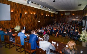 A imagem mostra um auditório com paredes marrons e uma plateia formada por homens e mulheres. Em primeiro plano está uma mesa solene de madeira também composta por homens e mulheres