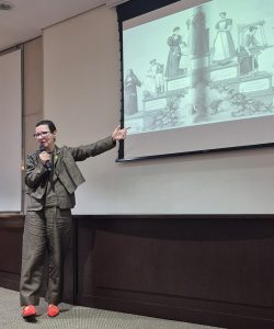 Legenda alternativa: A foto mostra um momento da palestra juíza Daniela de Freitas Marques, que tem cabelos curtos e usa óculos, vestindo um conjunto de calças e jaqueta cor cobre com sapatos laranja. Ela fala ao microfone e aponta para o telão onde está projetada uma obra que retrata, em preto e branco, o papel social da mulher no século XIX