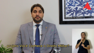 A imagem mostra o juiz de Direito Marcos Luiz Nery Filho sentado, de terno cinza, camisa branca e gravata azul. Ao seu lado direito há um quadro na parede com detalhes em azul e branco. No canto direito inferior da imagem está a janela da intérprete de libras, de camisa preta
