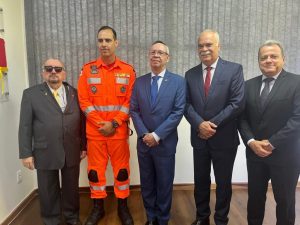 A imagem mostra o novo comandante do 2º BBM vestindo traje laranja dos bombeiros de Minas Gerais. Ele está em uma sala acompanhado por quatro representes do TJMMG. À esquerda está o presidente desembargador Jadir Silva usando terno preto, camisa branca, gravata amarela e óculos escuros. À direita está o desembargador Osmar Duarte Marcelino vestindo terno azul, gravata da mesma cor, camisa branca e usando óculos de grau; o desembargador Rúbio Paulino Coelho usando terno azul, camisa branca e gravata vermelha; e o chefe de gabinete coronel Giovanne Gomes da Silva, de terno e gravata pretos, e camisa branca. Ao fundo há uma parede branca e janela ampla com persiana cinza