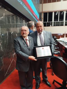 A imagem mostra o presidente do TJMMG, desembargador Jadir Silva, usando terno preto, gravata listrada e óculos. Ao lado está o professor Hermes Vilchez Guerrero vestindo terno cinza e gravata listrada. Os dois seguram a placa de alumínio com o título de Cidadão Honorário de Minas Gerais. Ao fundo da foto há uma parede branca com janelas e do lado esquerdo há uma parede de mármore preto e o telão do Plenário da ALMG