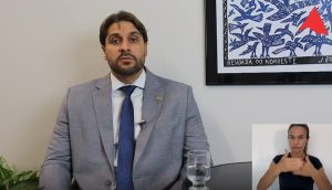A imagem mostra o juiz de Direito Marcos Luiz Nery Filho sentado, de terno cinza, camisa branca e gravata azul. Ao seu lado direito há um quadro na parede com detalhes em azul e branco. No canto direito inferior da imagem está a janela da intérprete de libras, de camisa preta
