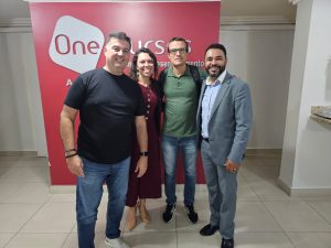 A imagem mostra um grupo de três homens e uma mulher. Eles estão de pé diante de um banner vermelho. O homem mais à direita da imagem é o professor Diocesio Sant'anna da Silva, de terno cinza