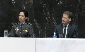 A imagem mostra uma mulher e um homem, sentados na tribuna da Assembleia Legislativa de Minas Gerais. O homem à direita é o desembargador Fernando Antônio Nogueira Galvão da Rocha, vestindo terno e gravata preta, e camisa branca. A mulher à esquerda é a comandante-geral do Corpo de Bombeiros Militar de Minas Gerais, coronel Jordana Daldegan, vestindo farda da corporação. Eles estão sentados atrás de uma mesa branca de mármore. A parede ao fundo é de mármore preto