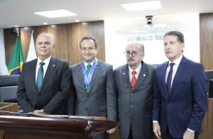 A imagem mostra quatro desembargadores na sala do Tribunal Pleno do TJMMG. À esquerda está o segundo vice-presidente do TJMG Saulo Versiani Penna, trajando um terno preto com gravata azul, seguido pelo presidente do TJMG Luiz Carlos de Azevedo Corrêa Junior, com terno cinza e gravata verde. A terceira pessoa é o presidente do TJMMG Jadir Silva, vestindo terno cinza e gravata vermelha, e por último está o diretor da EJM Fernando Antônio Nogueira Galvão da Rocha, com terno azul e gravata roxa