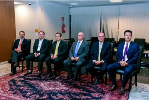 A foto mostra um dispositivo de honra formado por seis homens vestindo terno, sentados lado a lado em cadeiras pretas em um hall. O ambiente tem piso de mármore e um grande tapete de tons vermelho e azul. Ao fundo há poltronas pretas e uma parede com cortinas claras. O desembargador Fernando José Armando Ribeiro está à direita, de terno azul e gravata roxa. Ao seu lado estão os desembargadores José Arthur de Carvalho Pereira Filho, de gravata amarela; Saulo Versiani Penna, de gravata azul; Luiz Carlos de Azevedo Corrêa Junior, de gravata amarela; Adriano Bernardes de Souza, de camisa branca; e Pedro Carlos Bitencourt Marcondes, de gravata laranja