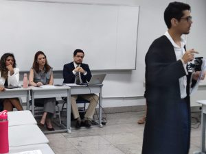 A imagem mostra homens e mulheres em uma sala, simulando um tribunal do júri. Ao centro há um homem vestindo toga. Ele fala para a plateia que inclui o juiz George Walter Barreto Paviotti, que usa terno azul, gravata listrada e calça bege