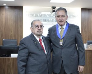 A foto mostra o presidente do TJMMG e o ministro do STJ na sala do Tribunal Pleno. O presidente do TJMMG veste terno cinza, camisa branca e gravata vermelha, usa óculos e tem outra armação pendurada por uma cordinha ao peito. O ministro também veste terno cinza, camisa branca, gravata com tons dourados e usa o Colar do Mérito Judiciário Militar, composto por uma medalha presa ao pescoço por uma fita azul. Eles estão diante de uma tribuna de madeira, e ao fundo há um painel com o nome do TJMMG por extenso