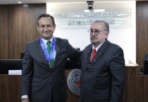 A foto mostra dois desembargadores lado a lado na sala do Tribunal Pleno do TJMMG. À esquerda está o presidente do TJMG Luiz Carlos de Azevedo Corrêa Junior, vestindo terno cinza e gravata verde, e à direita o presidente do TJMMG Jadir Silva, com terno preto, gravata vermelha e óculos. Ao fundo há um letreiro com o nome do TJMMG