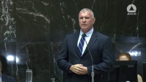 A imagem mostra o secretário de Estado de Justiça e Segurança Pública do Estado de Minas Gerais, Rogério Greco, vestindo terno preto e gravata listrada, falando ao microfone. A parede ao fundo é de mármore preto