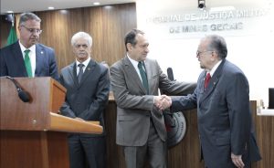 A imagem mostra quatro desembargadores na sala do Tribunal Pleno do TJMMG. À esquerda está o Corregedor-geral de Justiça, trajando um terno azul, camisa branca e gravata verde; seguido pelo corregedor da Justiça Militar, de terno e gravata cinza, e camisa branca. Eles olham o cumprimento entre o presidente do TJMG Luiz Carlos de Azevedo Corrêa Junior, com terno cinza e gravata verde, e o presidente do TJMMG Jadir Silva, vestindo terno cinza e gravata vermelha. Na frente deles há uma tribuna de madeira, e ao fundo o letreiro com o nome do TJMMG por extenso.