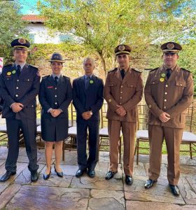 A imagem mostra cinco pessoas em um pátio aberto, com as mãos cruzadas à frente do corpo, sendo quatro homens e uma mulher. Os dois mais à esquerda da imagem são o coronel Moisés Magalhães de Sousa, chefe do Estado-Maior do CBMMG, e a coronel Jordana de Oliveira Filgueiras Daldegan, comandante-geral do CBMMG, vestindo farda oficial da corporação. Ao centro está o desembargador Sócrates Edgard dos Anjos, usando terno, camisa e gravata cinzas. À direita da imagem estão o coronel Frederico Otoni Garcia, comandante-geral da PMMG, e o coronel Maurício José de Oliveira, chefe do Estado-Maior da PMMG, ambos vestindo farda oficial da PMMG.