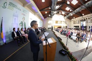Imagem mostra o Centro de Artes e Convenções da Ufop, com o governador Romeu Zema fazendo discurso em um púlpito de madeira com microfone, diante de diversas autoridades entre homens e mulheres na tribuna de honra, e uma plateia com diversos convidados