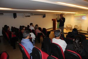 Imagem mostra o auditório do TJMMG, com um público em primeiro plano formado por homens e mulheres sentados na plateia. Eles assistem à palestra do servidor Gustavo Cândido, um homem de camisa escura, óculos e barba. Ao fundo há um telão escrito desregulação emocional