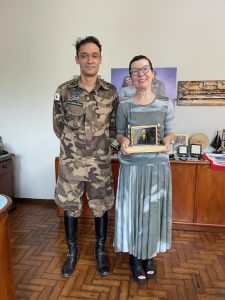 A foto mostra a juíza Daniela de Freitas Marques segurando um troféu em madeira com sua foto aplicada. Ela é uma mulher de cabelos curtos, está de vestido em tons de cinza e sapato preto. Ao seu lado está o major Jordane César, com farda da cavalaria da Polícia Militar de Minas Gerais e botas de cavalgar. Eles estão em uma sala onde, ao fundo, há uma aparador de madeira com fotos, troféus e medalhas