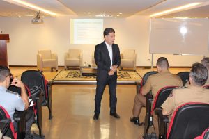 A imagem mostra o auditório do TJMMG. Em destaque está o desembargador Fernando Antônio Nogueira Galvão da Rocha, de terno escuro e camisa branca. Ele fala para uma plateia de policiais militares e bombeiros militares