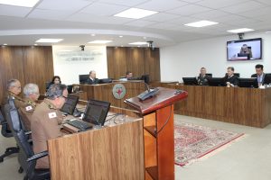 A foto mostra a composição do Tribunal Pleno. Do lado esquerdo há uma tribuna com três desembargadores fardados com roupa da PMMG. Ao centro, uma mulher e dois homens, um deles o presidente que presidiu a sessão vestindo toga. Na tribuna direita, três homens, dois deles desembargadores vestindo toga, e um com fardamento do Corpo de Bombeiros. Ao fundo há uma placa com a logomarca do TJMMG, composta por um triângulo estilizado da bandeira de Minas Gerais e o nome do tribunal por extenso