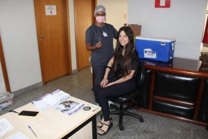 A foto mostra a colaboradora Mayni Grisolia Gualberto de Oliveira sentada enquanto recebe a vacina. Ela está sorrindo e usa blusa e calça pretas. A profissional que aplica a vacina usa traje cinza, máscara e touca. Ao fundo há paredes brancas, portas de madeira e uma estante com uma caixa térmica azul onde estão acondicionadas as vacinas