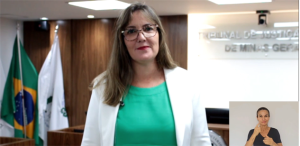 A imagem mostra a coordenadora da Gestão Documental vestindo blusa verde e blaser branco. Ela é uma mulher branca, de óculos e cabelos compridos. Ela está na sala do Tribunal Pleno, com duas bandeiras no fundo e o nome do tribunal em um letreiro. No canto direito da imagem está a janela da intérprete de libras, de camiseta preta e cabelo preso 
