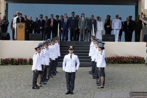 A imagem mostra um grupo de homens e mulheres em cima de um palco montado na Praça Tiradentes, em Ouro Preto, tendo à frente um destacamento da Polícia Militar de Minas Gerais vestindo farda branca da corporação